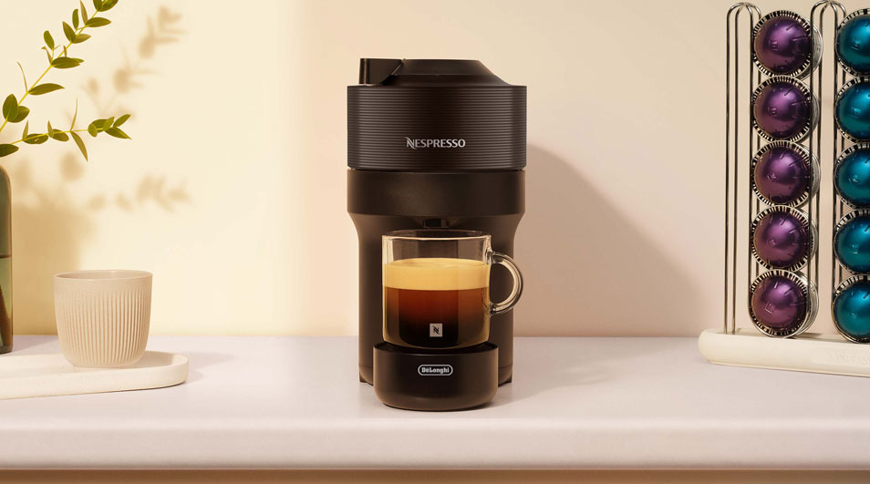 Zwarte Nespresso-koffiemachine met een glazen kopje espresso eronder, naast een kopjeshouder met kleurrijke capsules op een lichte keukenondergrond.
