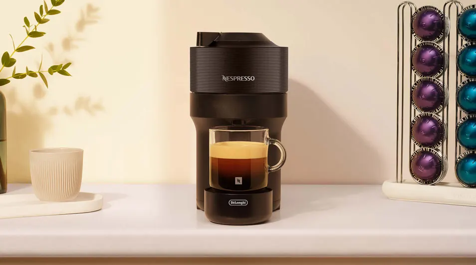 Zwarte Nespresso-koffiemachine met een glazen kopje espresso eronder, naast een kopjeshouder met kleurrijke capsules op een lichte keukenondergrond.