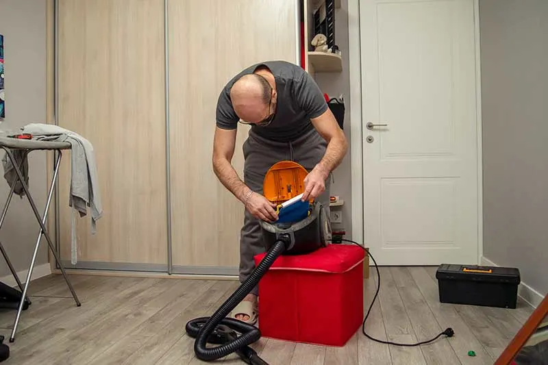 Man in een kamer plaatst een nieuwe stofzuigerzak in een sledestofzuiger die op een rode poef staat.