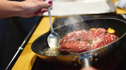 Een zwarte pan op een kookplaat bevat vlees dat wordt gekookt, met een zilveren lepel die wordt gebruikt om de olie te verdelen.