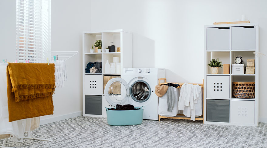 Een wasruimte met een open wasmachine, een blauwe wasmand, rekken en een droogrek tegen een witte muur.