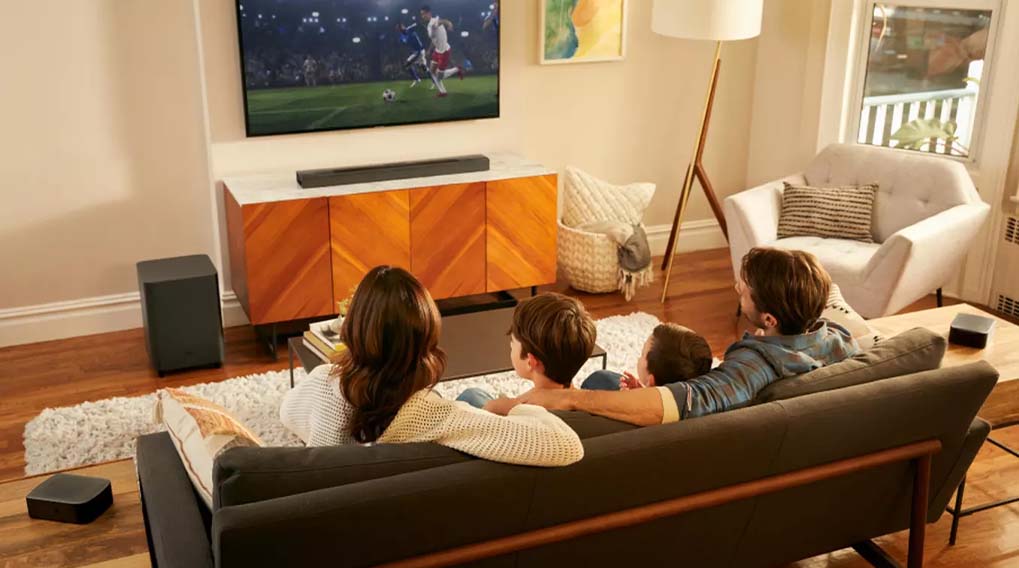 Een familie kijkt tv in een woonkamer met een kast, een lamp, een stoel en een grijze bank.