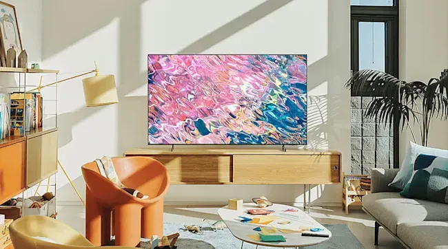 In een zonnige woonkamer staat een tv op een houten meubel; een oranje stoel, lage tafel en grijze bank omringen de ruimte.