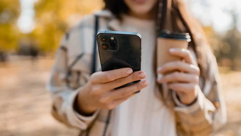 Vrouw houdt een zwarte smartphone en een bruine kartonnen beker vast. De achtergrond is herfstachtig.