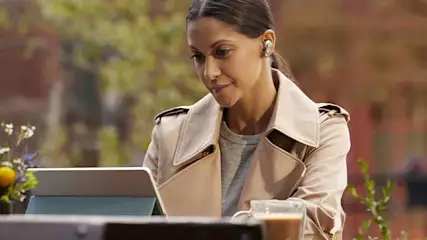 Vrouw met oortjes, beige jas, grijze trui kijkt naar een tablet. Koffie en bloemen op tafel, buitenlocatie met bomen op achtergrond.