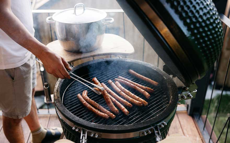 Sausages cook on a round barbecue as someone turns them with tongs, surrounded by smoke and a cosy outdoor feel.