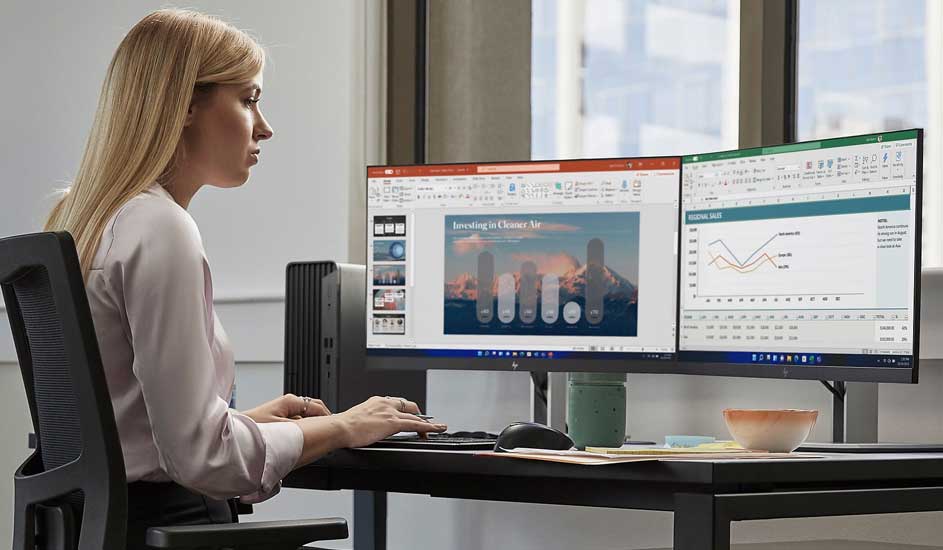 Woman working intently at a desk with dual wide monitors displaying charts and slides in a calm modern office space.