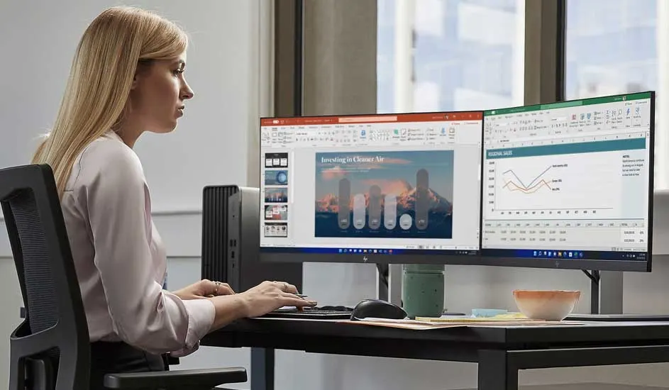 Woman working intently at a desk with dual wide monitors displaying charts and slides in a calm modern office space.