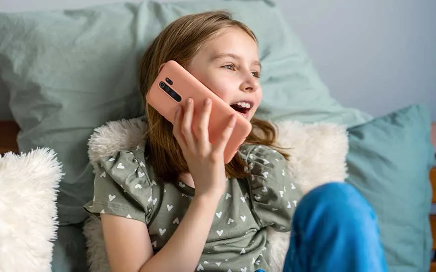 A girl relaxes among fluffy cushions on her bed, holding a large smartphone to her ear while chatting happily.