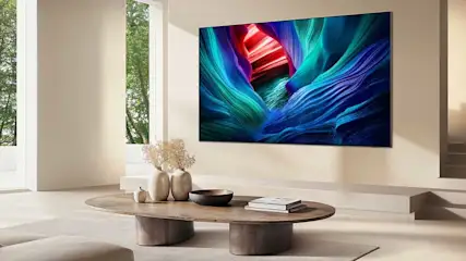 Een moderne woonkamer met grote tv aan de muur toont levendige blauwe en rode grafische inhoud. Minimalistisch design met houten tafel, bloemenvazen en natuurlijk licht van ramen.