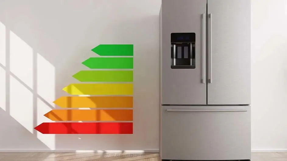 A refrigerator stands next to a wall with an energy efficiency label. The label has arrows colored green, yellow, orange, and red.