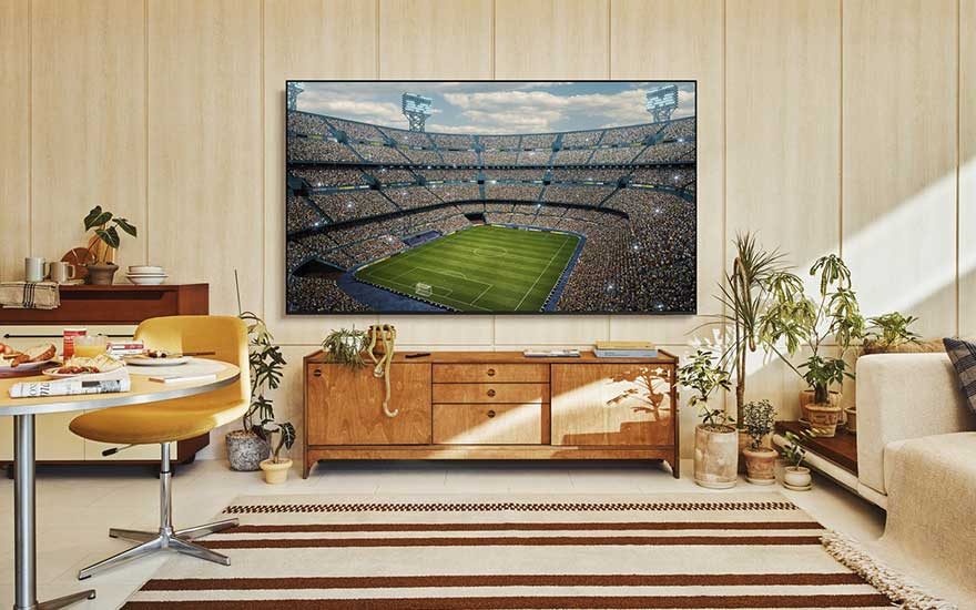 Gezellige woonkamer met retro-meubels, veel planten en een muur-tv die een overvol voetbalstadion tijdens een wedstrijd toont.