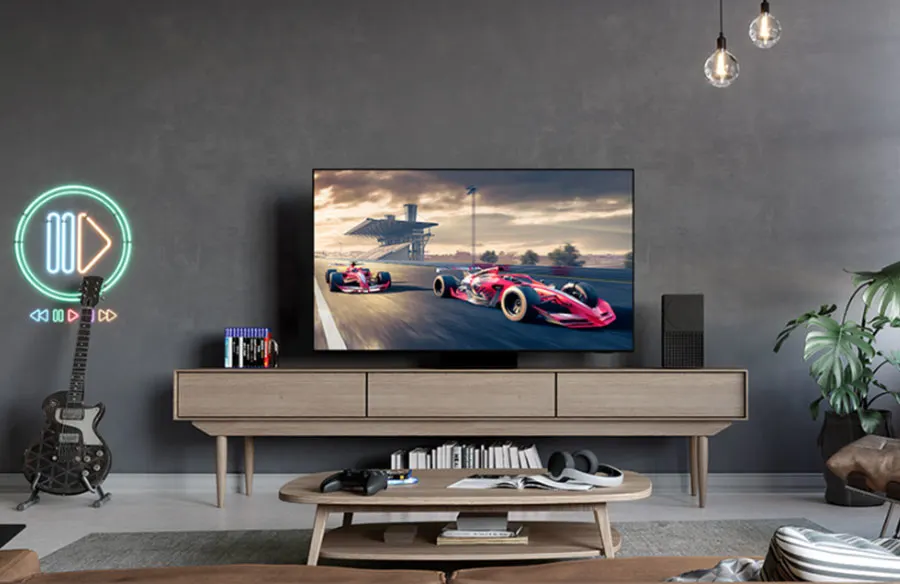 Je ziet een Samsung-televisie op een lichtbruine tv-kast staan in een woonkamer setting. De muur is donkergrijs. Voor de tv-kast staat een ovalen lichtbruine salontafel en je ziet een klein stukje van een donkerbruine bank. Op de lichtgrijze vloer licht een grijs vloerkleed.