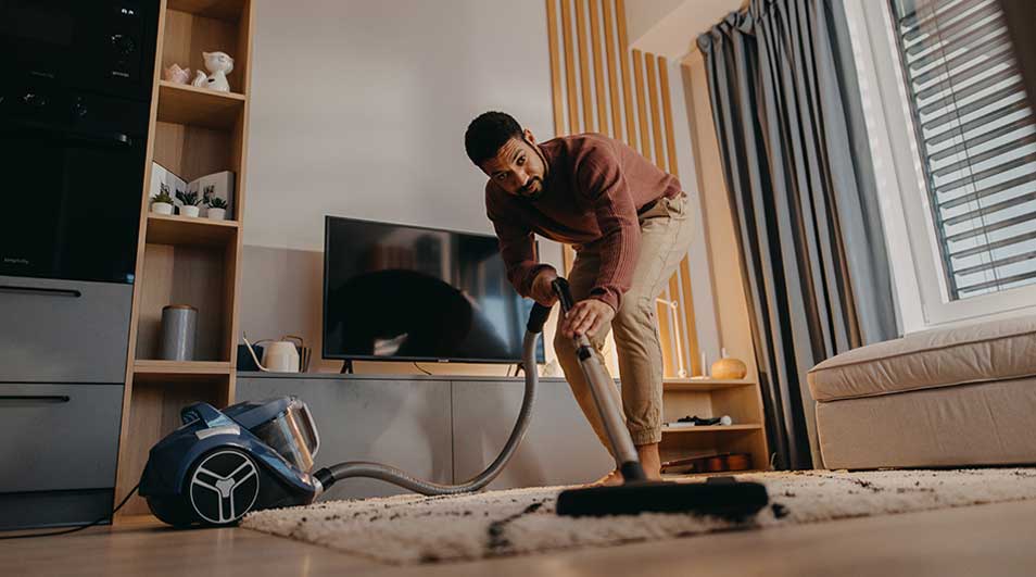 Een persoon stofzuigt zorgvuldig een licht vloerkleed in een moderne, warme woonkamer met tv-meubel, grote bank en zonlicht door het raam.