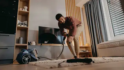 Een persoon stofzuigt zorgvuldig een licht vloerkleed in een moderne, warme woonkamer met tv-meubel, grote bank en zonlicht door het raam.