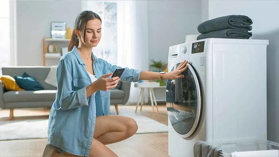 Een vrouw bedient met haar smartphone een moderne wasmachine in een lichte woonkamer. Op de wasmachine liggen opgevouwen handdoeken. Merk is niet zichtbaar.