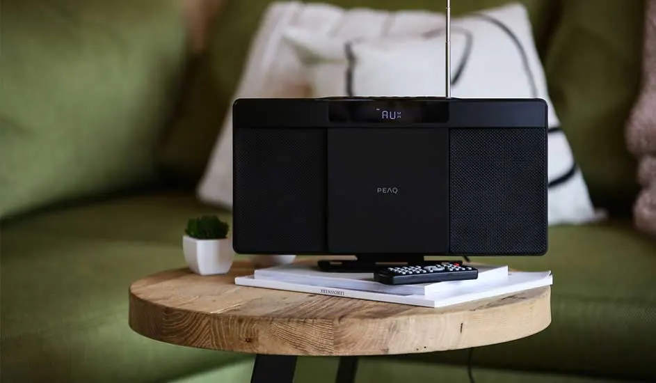 Houten wand achter een luidspreker op een bruine standaard. Een witte bank met decoratie en een bandrecorder staan links.