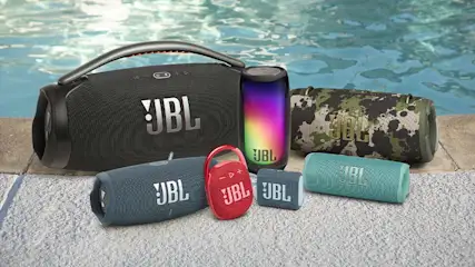Diverse JBL-luidsprekers staan op een betegelde rand bij een zwembad met blauw water. Zwart, camouflage, rood en regenboog kleuren.