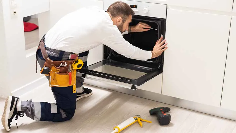 Een man met een gereedschapsriem repareert een witte oven in een witte keuken, met gereedschap op de vloer.