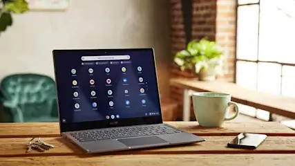 Laptop op houten tafel met sleutels, kopje en telefoon. Op de achtergrond een bakstenen muur met raam en een plant.