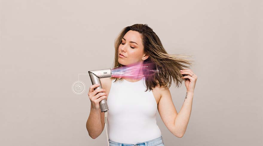 Vrouw gebruikt zilverkleurige föhn. Roze lichteffect om haar, tegen een lichtgrijze achtergrond. Ze draagt een wit hemdje en een blauwe jeans.