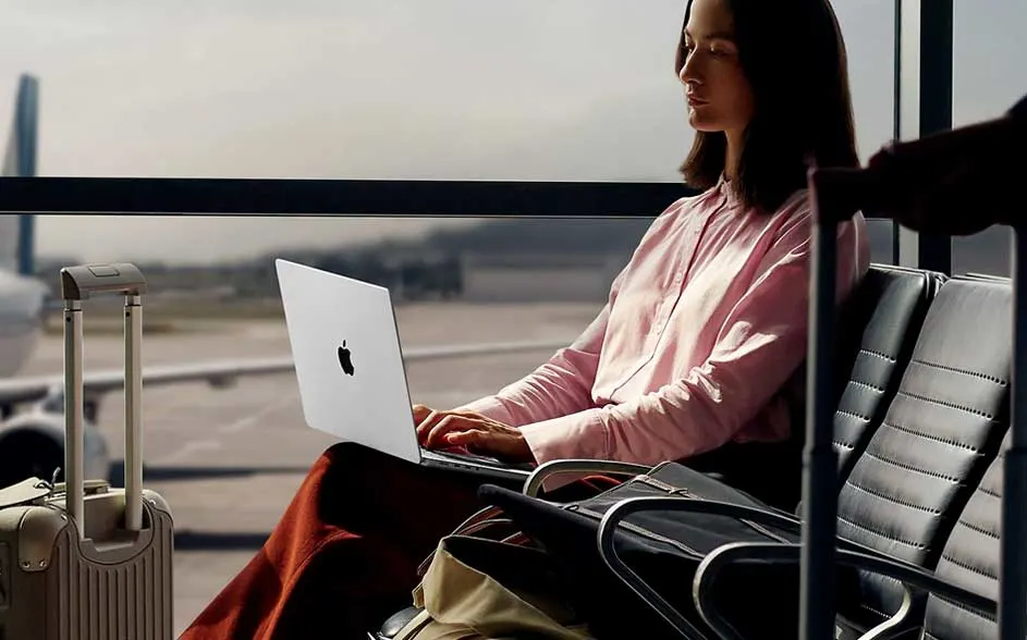 Een vrouw in een luchthavenlounge werkt op een zilveren Apple-laptop; naast haar staan koffers en tassen, met vliegtuigen buiten het raam.