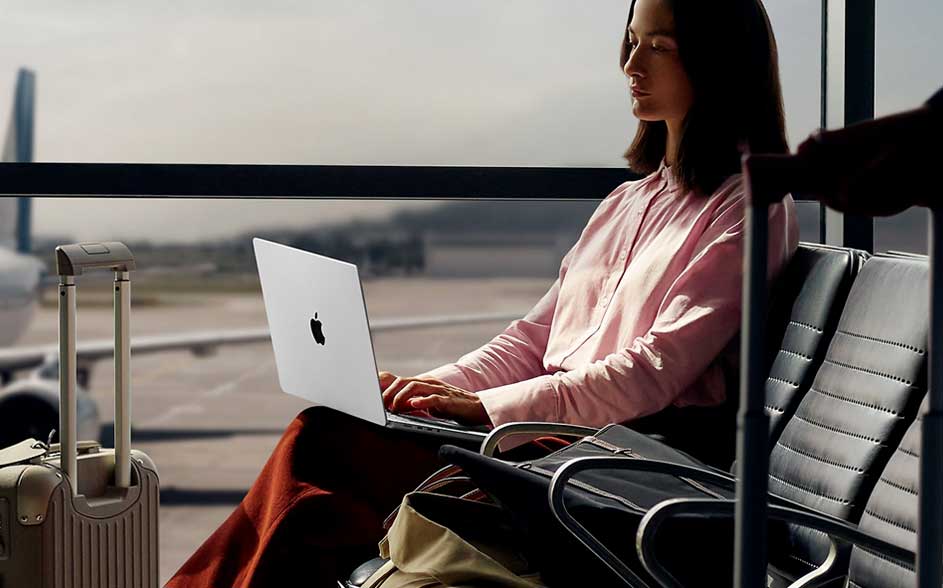 A woman in an airport lounge works on a silver Apple laptop; suitcases and bags sit nearby, with aircraft visible beyond the window.