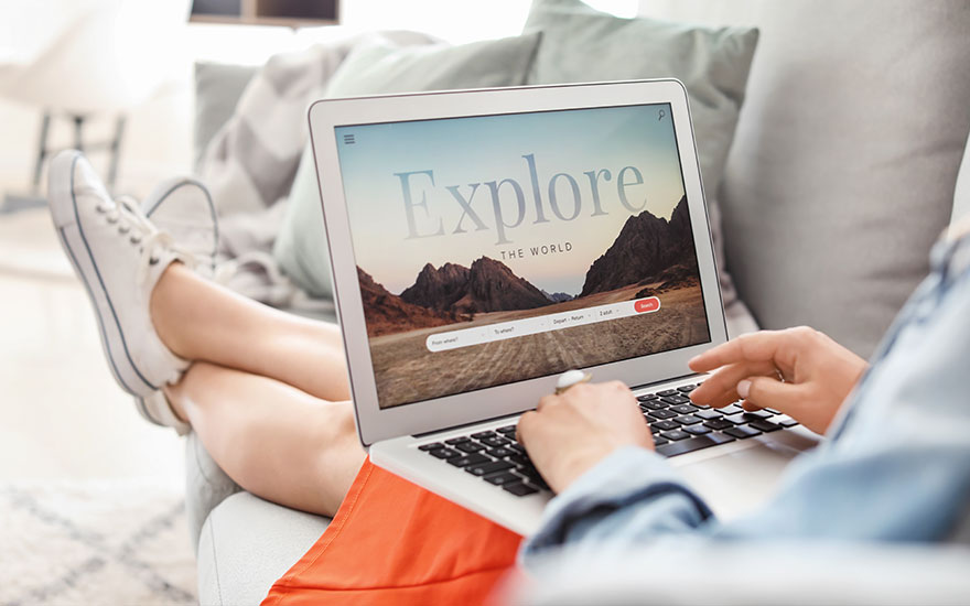 Persoon in oranje broek gebruikt laptop op bank, scherm toont "Explore the World" met berglandschap, witte sneakers, groene kussens.