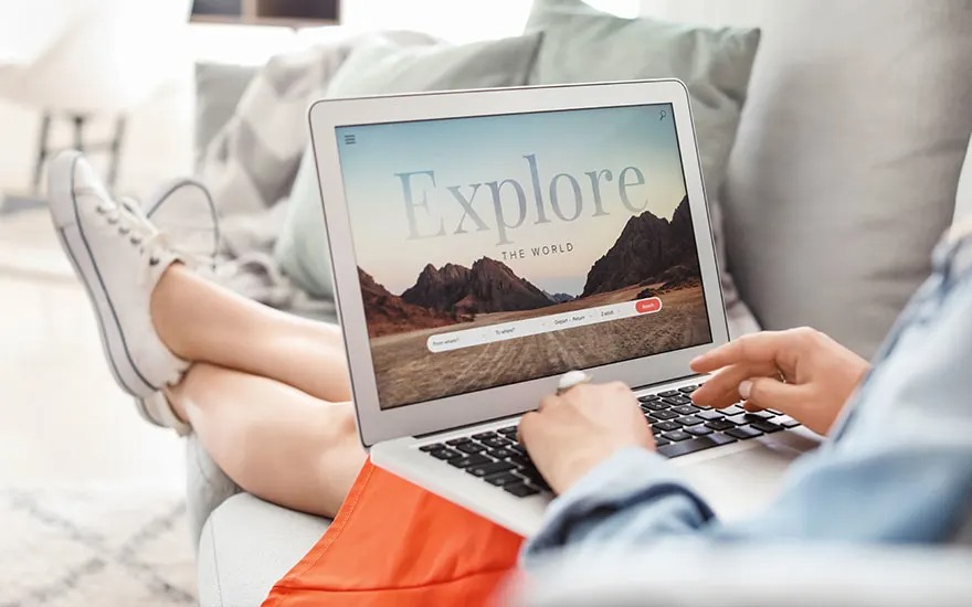 Person relaxing on sofa with feet up, browsing a travel site on laptop showing “Explore the world” over a mountain landscape.
