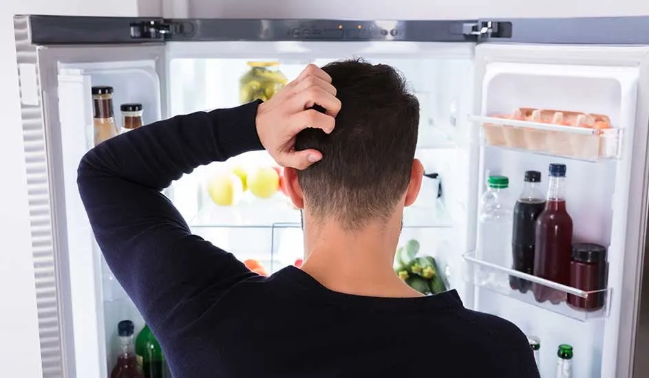 Een man staat peinzend voor een open Bosch-koelkast met scharniermechanisme, gevuld met eten en drinken in een moderne keuken.
