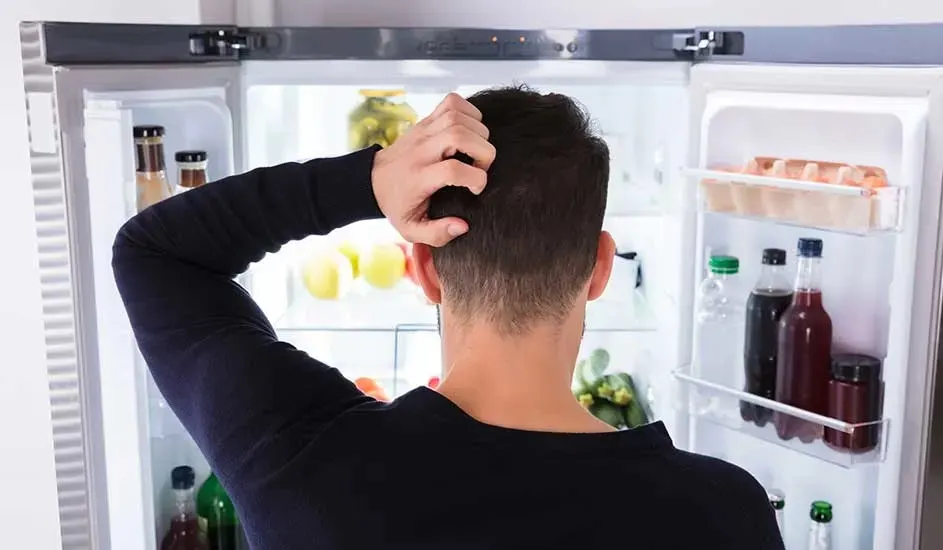 Een man staat peinzend voor een open Bosch-koelkast met scharniermechanisme, gevuld met eten en drinken in een moderne keuken.