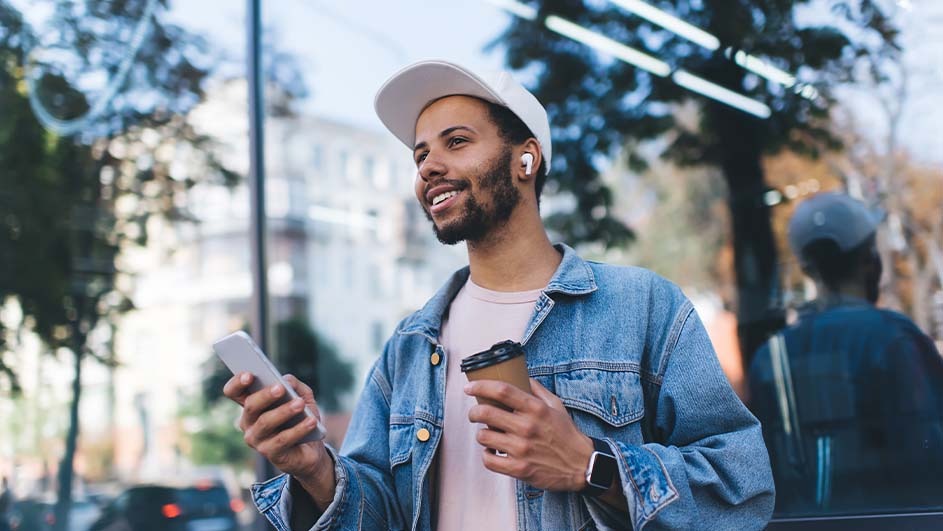 Man in a denim jacket holds a phone and coffee. He wears a cap and earbuds, standing near a building with trees visible.