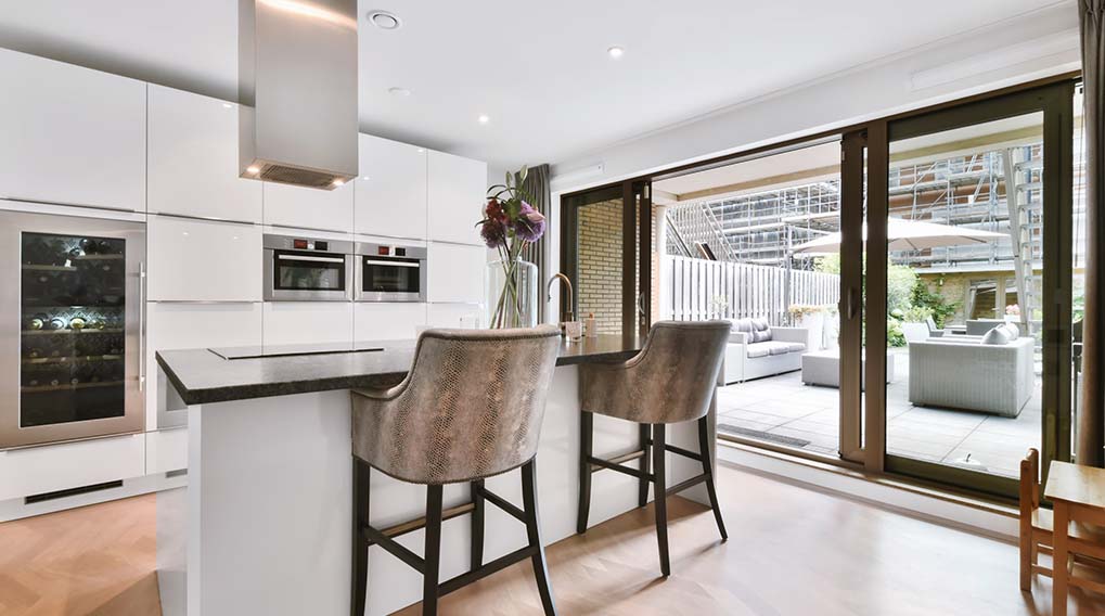 Een moderne keuken met witte kasten, een eiland met twee stoelen, en een schuifpui naar een terras met meubels.