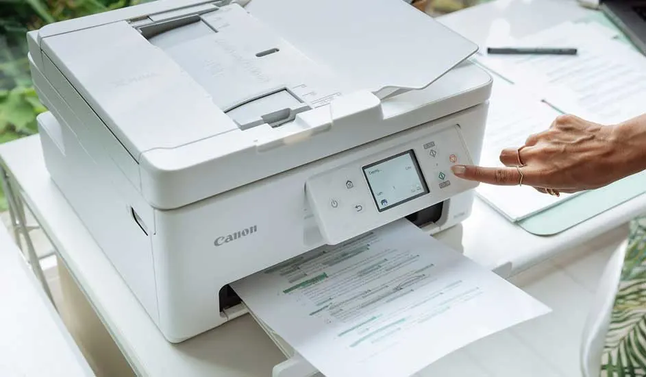 Een witte Canon‑printer staat op een bureau; een hand drukt op de Start‑knop terwijl een gemarkeerd document uit de lade wordt geprint.