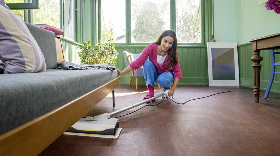 Woman uses a slim steam mop under the sofa in a bright living room, easily reaching hidden dust and dirt in tight, awkward spaces.
