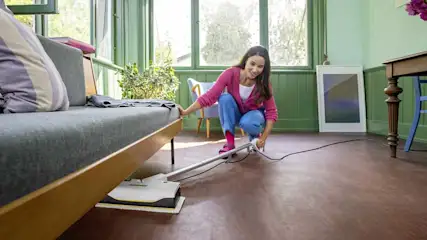 Woman uses a slim steam mop under the sofa in a bright living room, easily reaching hidden dust and dirt in tight, awkward spaces.