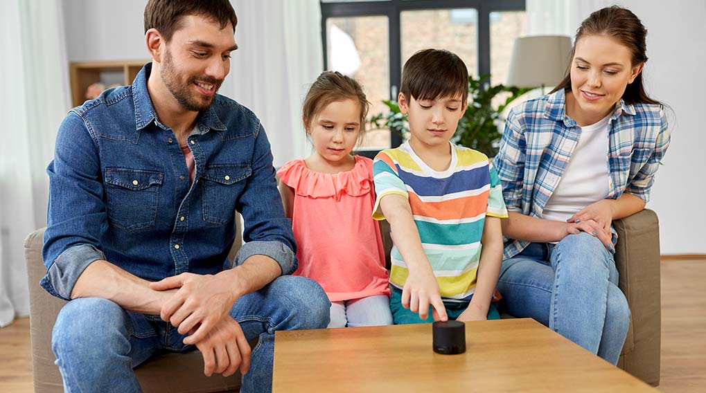Een gezin zit rond een houten tafel met een zwarte slimme speaker. Een kind raakt de speaker aan.