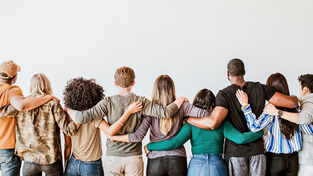 Van achteren gezien, omarmen negen mensen elkaar voor een witte achtergrond. Ze dragen diverse tops en broeken.