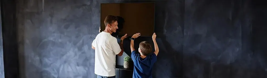 A man and a child hang a framed picture on a textured dark wall above a cabinet with a potted plant.