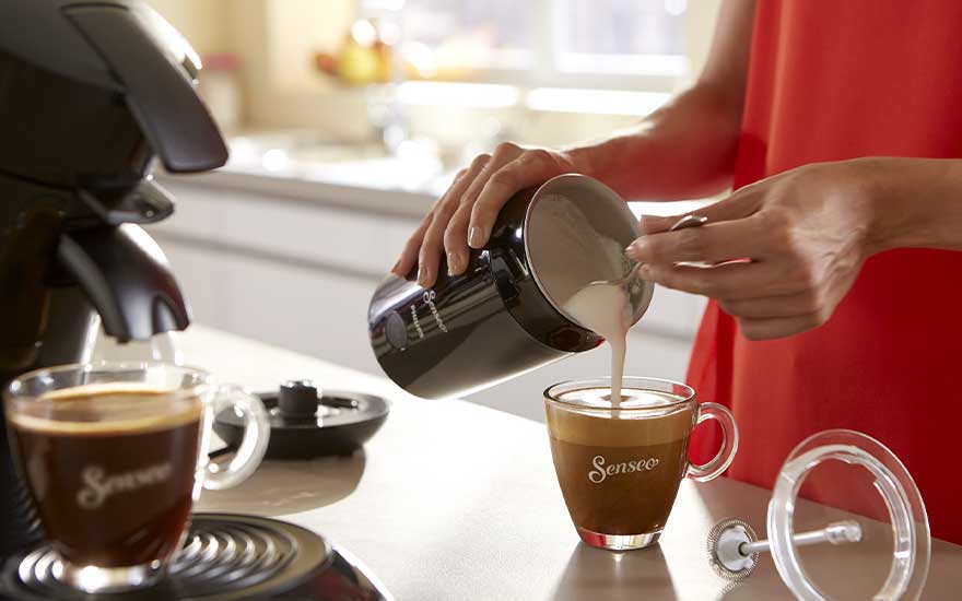 A person in a red dress pours creamy milk from a black jug into a Senseo coffee cup beside a pod coffee machine.