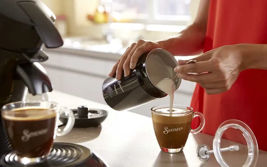 A person in a red dress pours creamy milk from a black jug into a Senseo coffee cup beside a pod coffee machine.