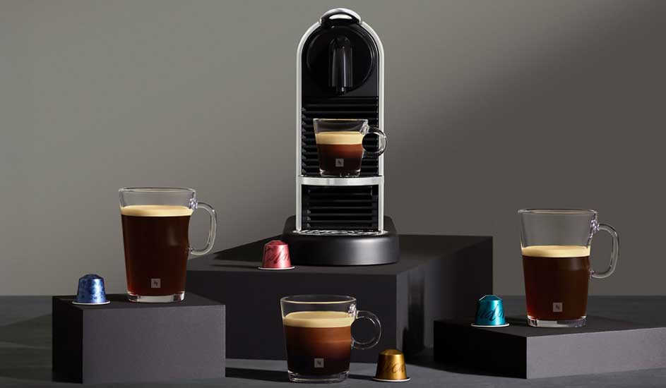 Een Nespresso-apparaat met drie glazen koffiekopjes en vier gekleurde capsules staat op zwarte blokken tegen een grijze achtergrond.
