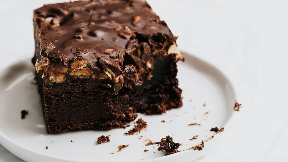 Een brownie met chocoladeglazuur en noten op een wit bord, met kruimels aan de zijkant.