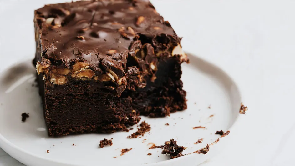 Een brownie met chocoladeglazuur en noten op een wit bord, met kruimels aan de zijkant.