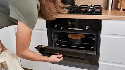 Vrouw opent zwarte oven in witte keuken; een taart staat op een rooster. Achter de oven is een zwart gasfornuis te zien.