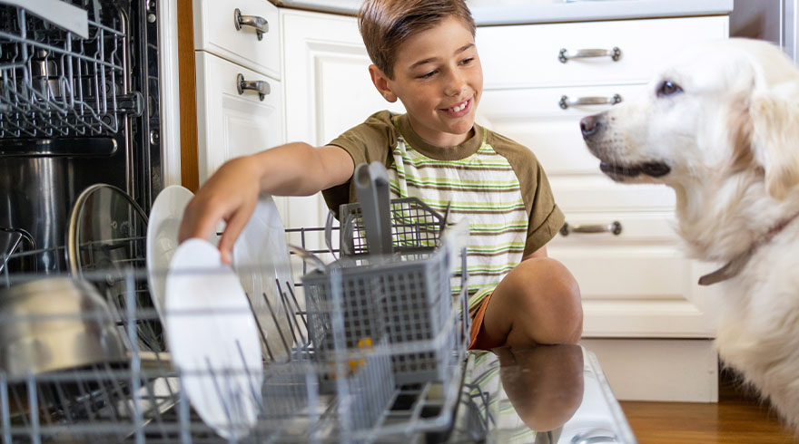 Jongen laadt witte borden in de vaatwasser, hond kijkt toe. Witte keukenkasten op achtergrond.