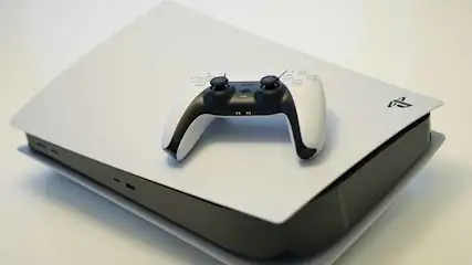 Een witte PlayStation 5 met een zwart-witte controller bovenop, op een lichte ondergrond. Het PlayStation logo staat rechtsboven.