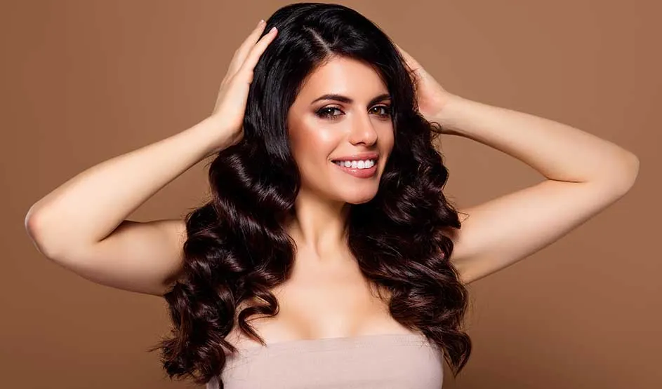 A smiling woman with long, dark, wavy hair holds her hands in her hair against a plain brown background.