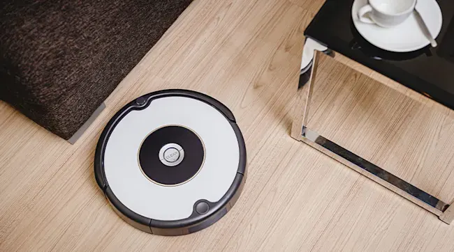 A robot vacuum sits on a wood floor near a dark sofa and a glass-topped table with a white cup and saucer.