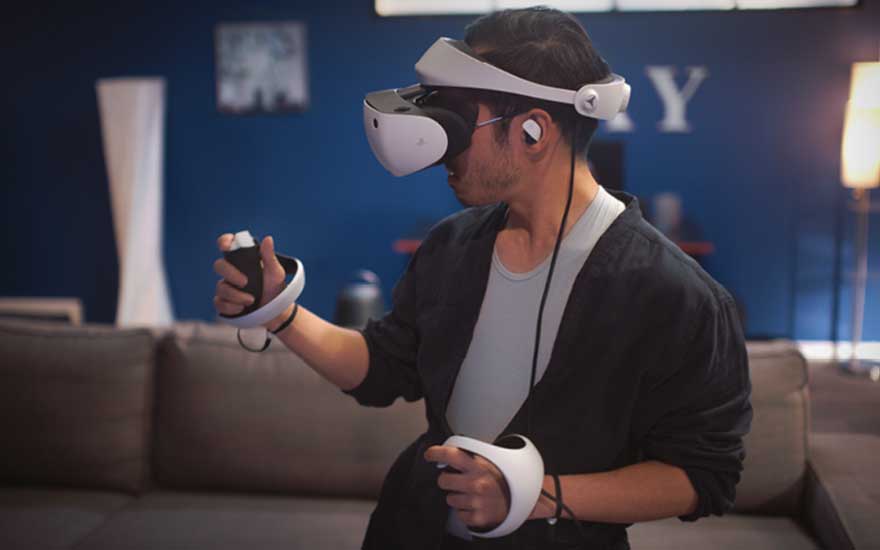 A gamer wearing a VR headset holds two motion controllers, standing before the sofa and fully immersed in a vivid virtual world.